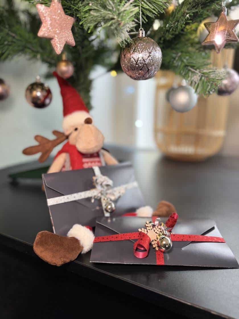 Zwei festlich verpackte Milanee Thaimassage Gutscheine liegen unter einem geschmückten Weihnachtsbaum, dekoriert mit roten Bändern, Glöckchen und winterlichen Details.