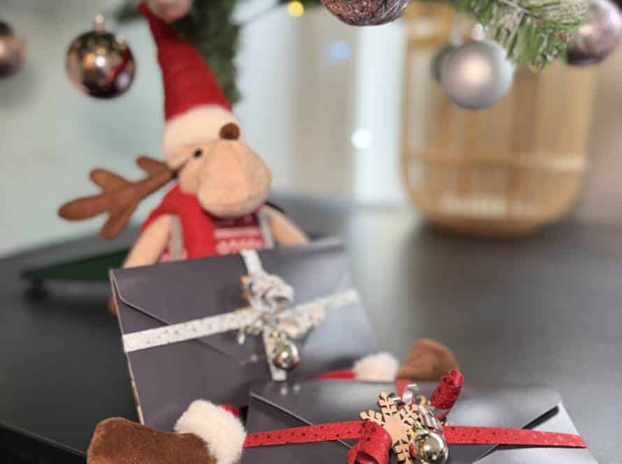 Zwei festlich verpackte Milanee Thaimassage Gutscheine liegen unter einem geschmückten Weihnachtsbaum, dekoriert mit roten Bändern, Glöckchen und winterlichen Details.
