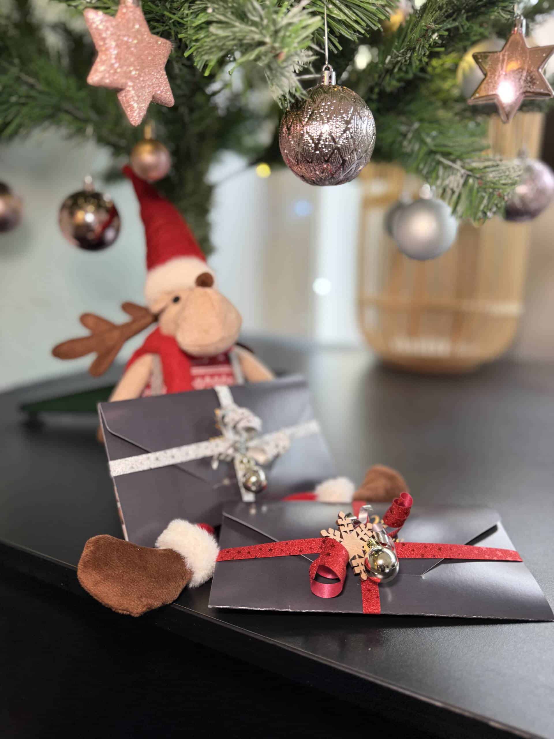 Zwei festlich verpackte Milanee Thaimassage Gutscheine liegen unter einem geschmückten Weihnachtsbaum, dekoriert mit roten Bändern, Glöckchen und winterlichen Details.