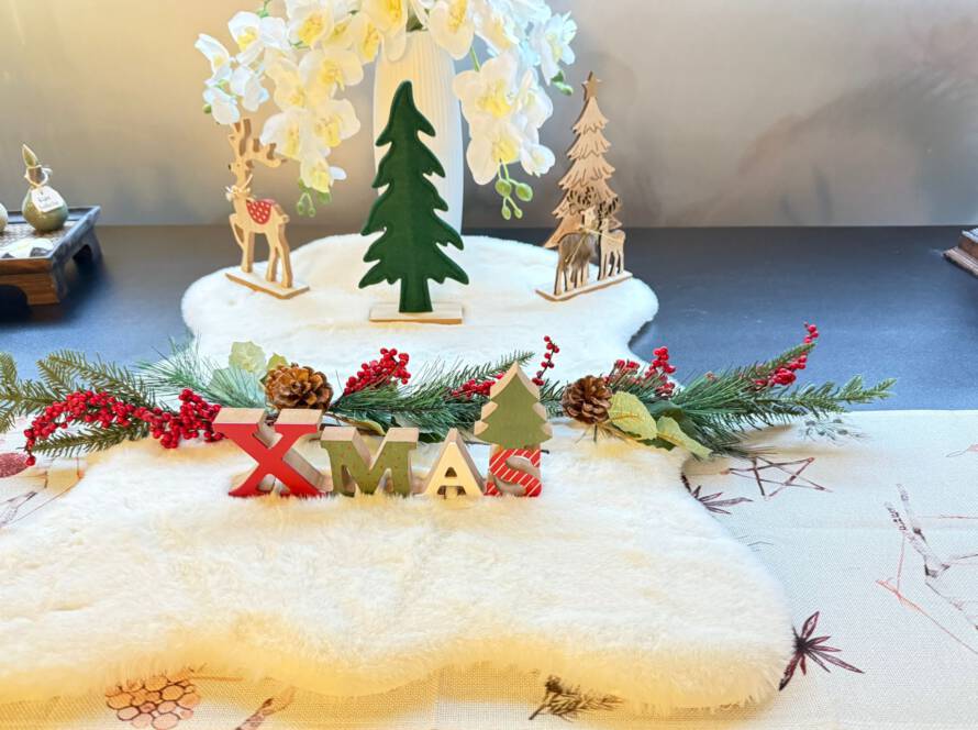 „Weihnachtliche Tischdekoration mit XMAS-Schriftzug, Tannenzweigen, Beeren und Holzfiguren im Hintergrund.“