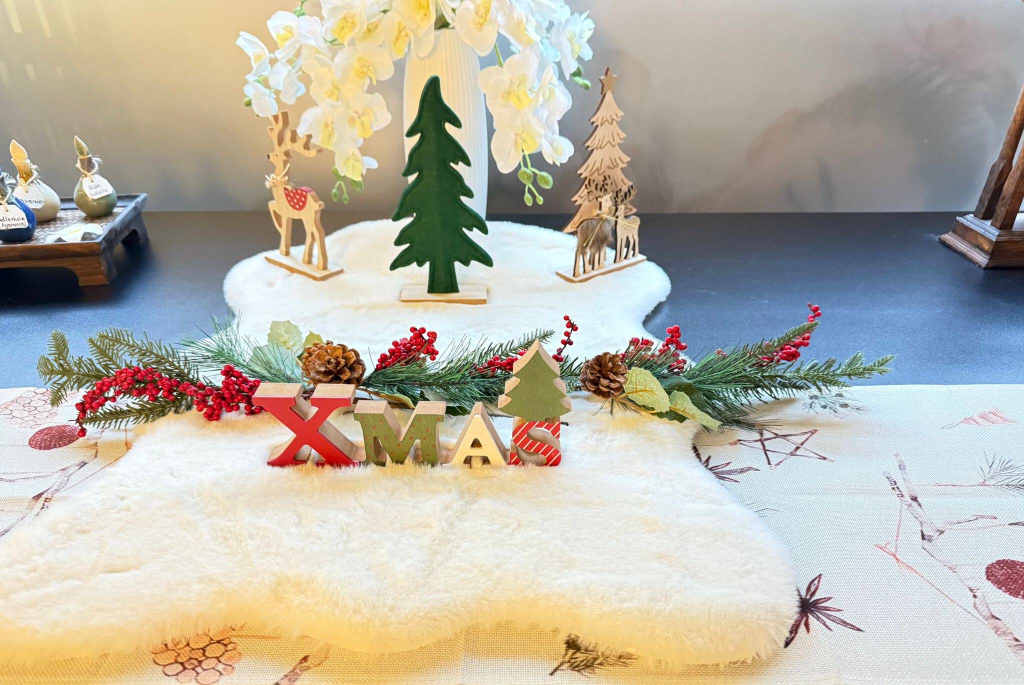 „Weihnachtliche Tischdekoration mit XMAS-Schriftzug, Tannenzweigen, Beeren und Holzfiguren im Hintergrund.“